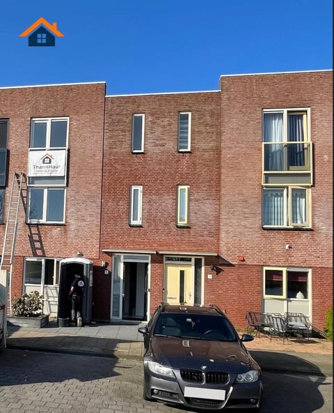 Red brick residential apartment building with multiple windows and a parked dark car in front on a sunny day