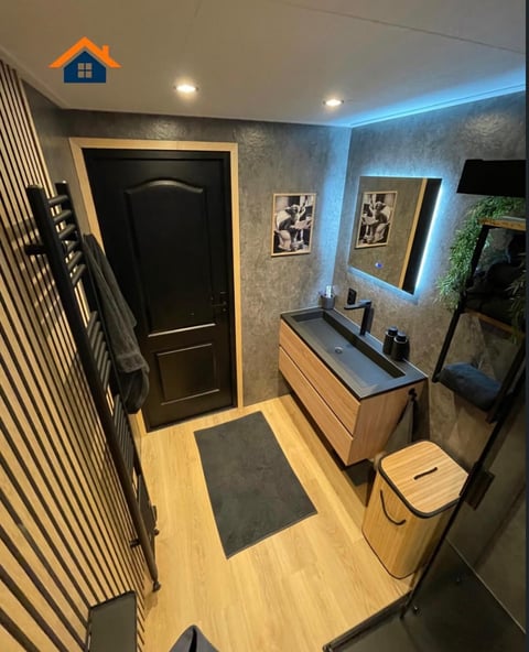 Modern bathroom with light wood flooring, blue accent lighting, gray wallpaper, wooden vanity with sink, black door, and framed artwork