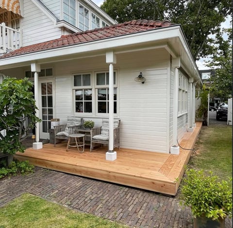 Modern white house extension with wooden deck, white support columns, and large windows overlooking a garden patio area