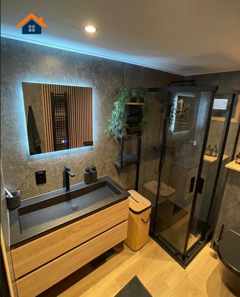 Modern bathroom with illuminated mirror, wooden vanity, black tiled shower enclosure, and decorative plants